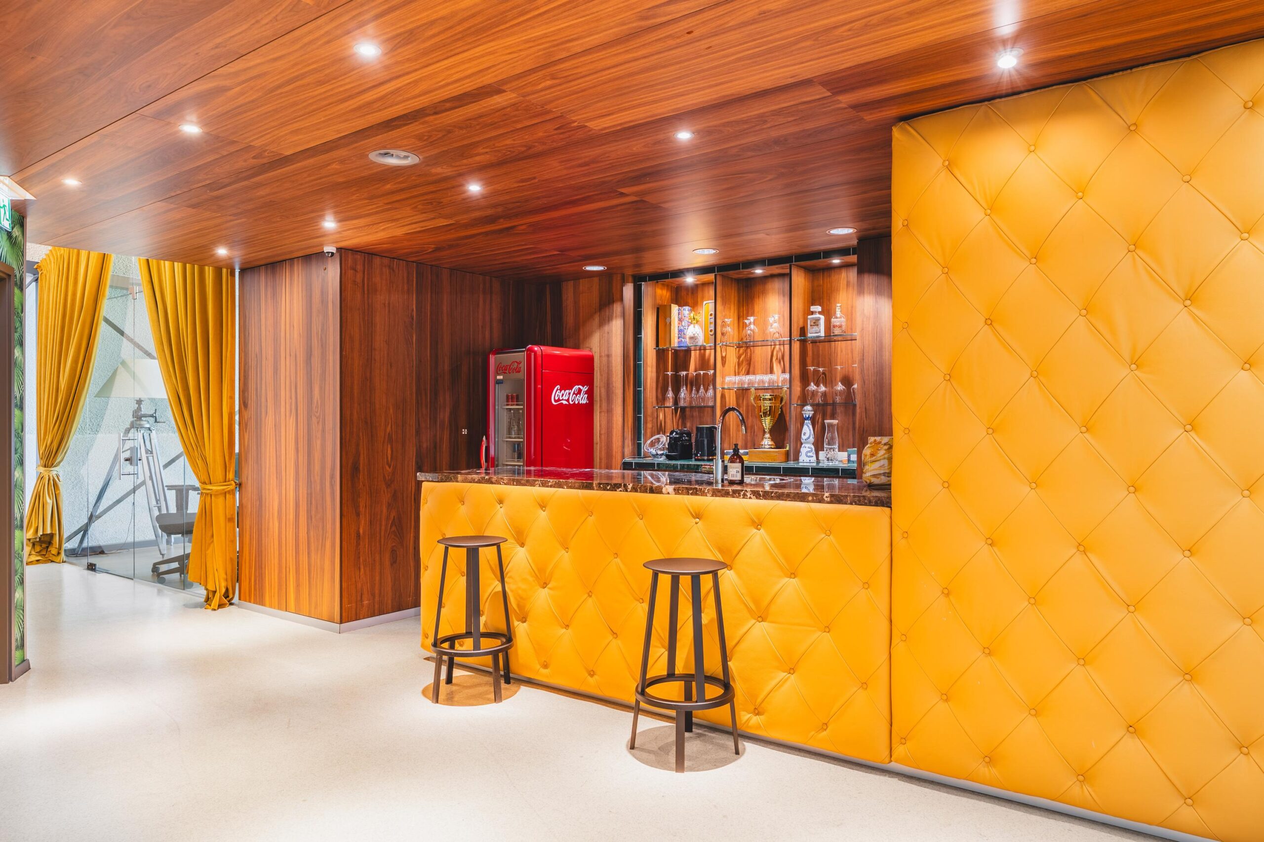 Moderne bar met gele, gewatteerde bekleding, houten accenten en een rode Coca-Cola koelkast aan de Prof. W.H. Keesomlaan.